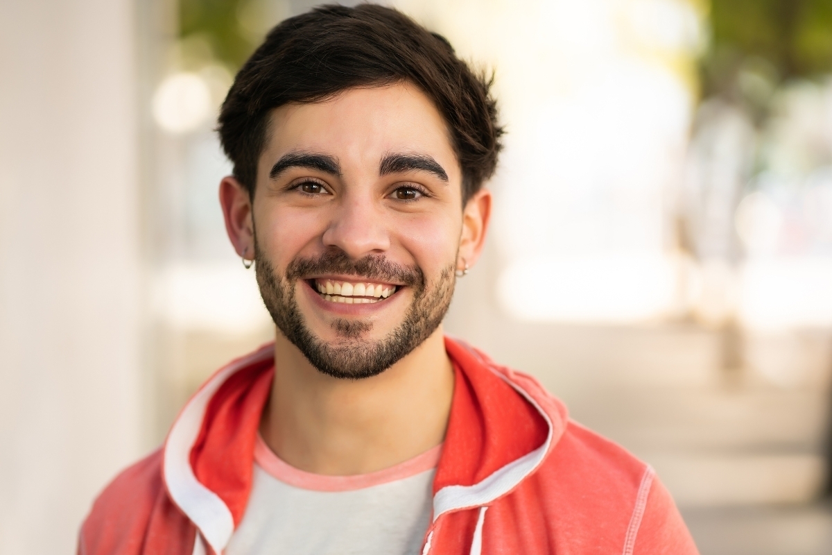 Smiling man with bright white teeth wearing a red hoodie in a dental office setting.